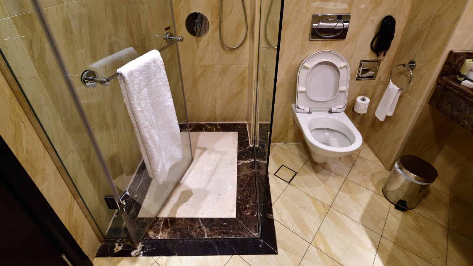 A modern bathroom with a glass shower, white towel, toilet, and beige marble tiles&mdash;perfect for guests over 35 booking a hotel seeking comfort and style.