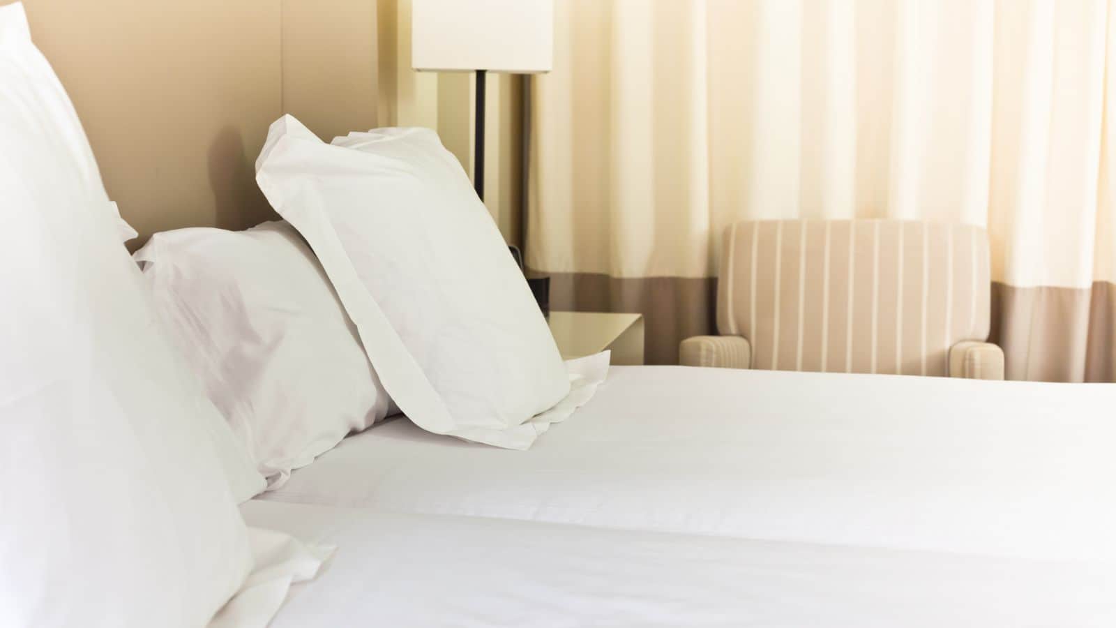 Two neatly made beds with white linens beside a nightstand, lamp, and a striped armchair in a hotel room&mdash;ideal for travelers seeking hotels for over 35 and looking for comfort during their stay.