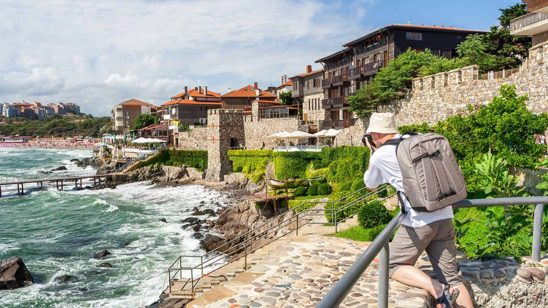 A person with a backpack photographs seaside stone buildings along a rocky coast on a sunny day.
