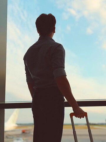 A person with luggage looks out airport window as a plane takes off into the sky.