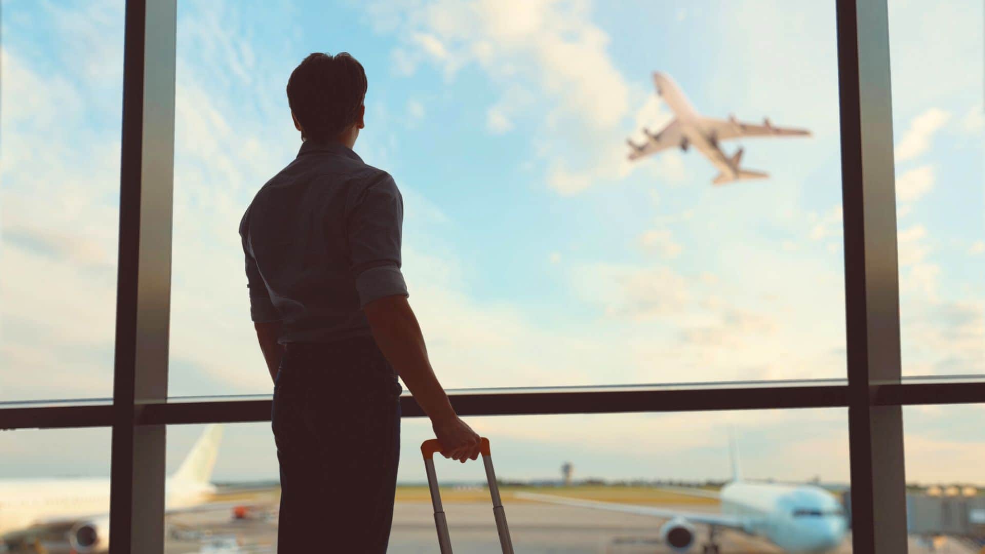 A person with luggage looks out airport window as a plane takes off into the sky.