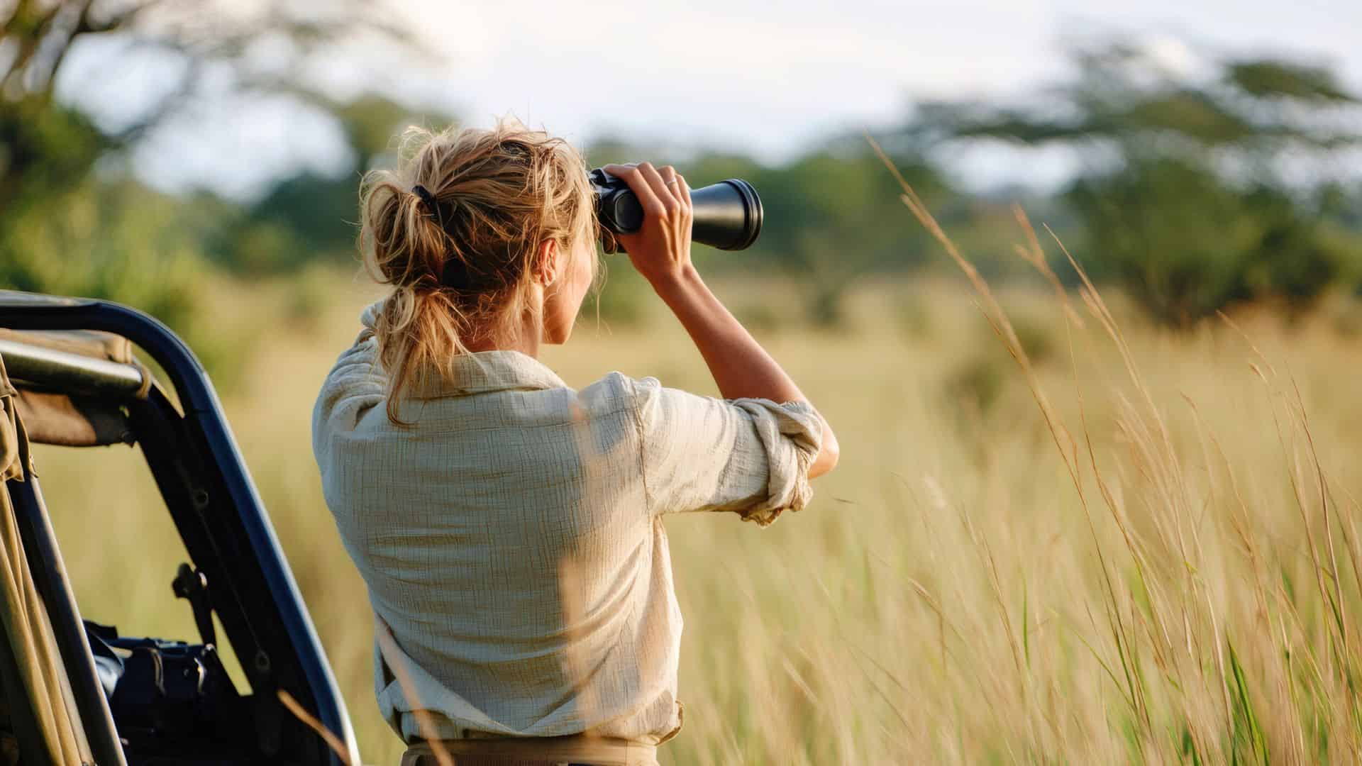 A person with binoculars observes wildlife from a safari vehicle in a grassy field.