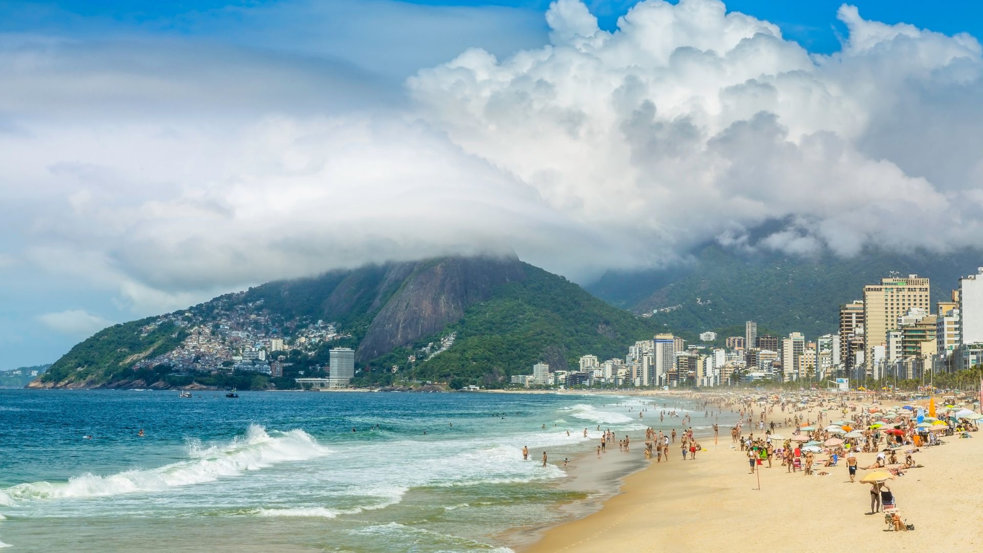 Crowded beach with waves, city buildings, and green mountains under a cloudy blue sky.