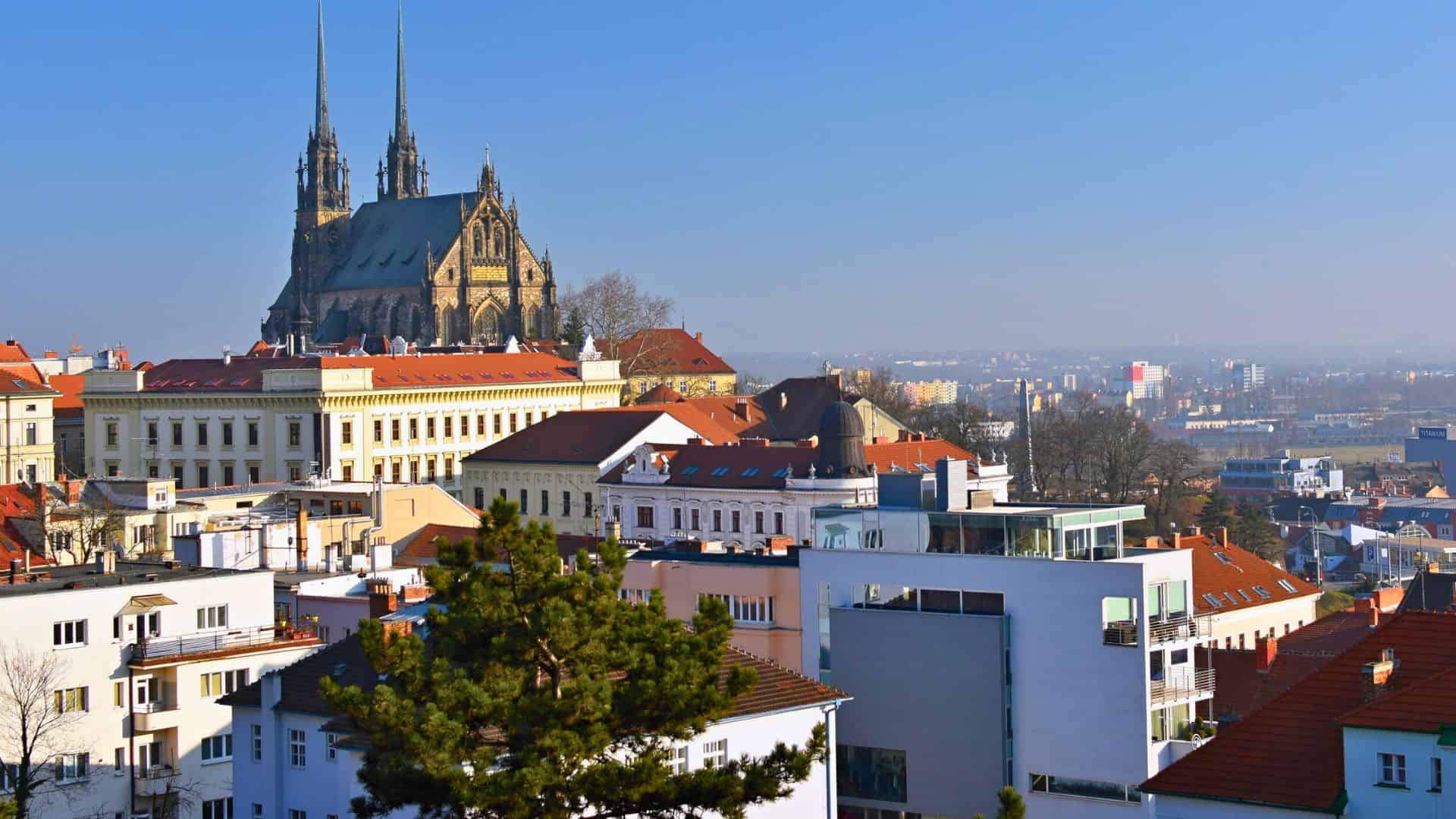 Cityscape with red-roofed buildings and a large cathedral with twin spires in the background under a clear sky.
