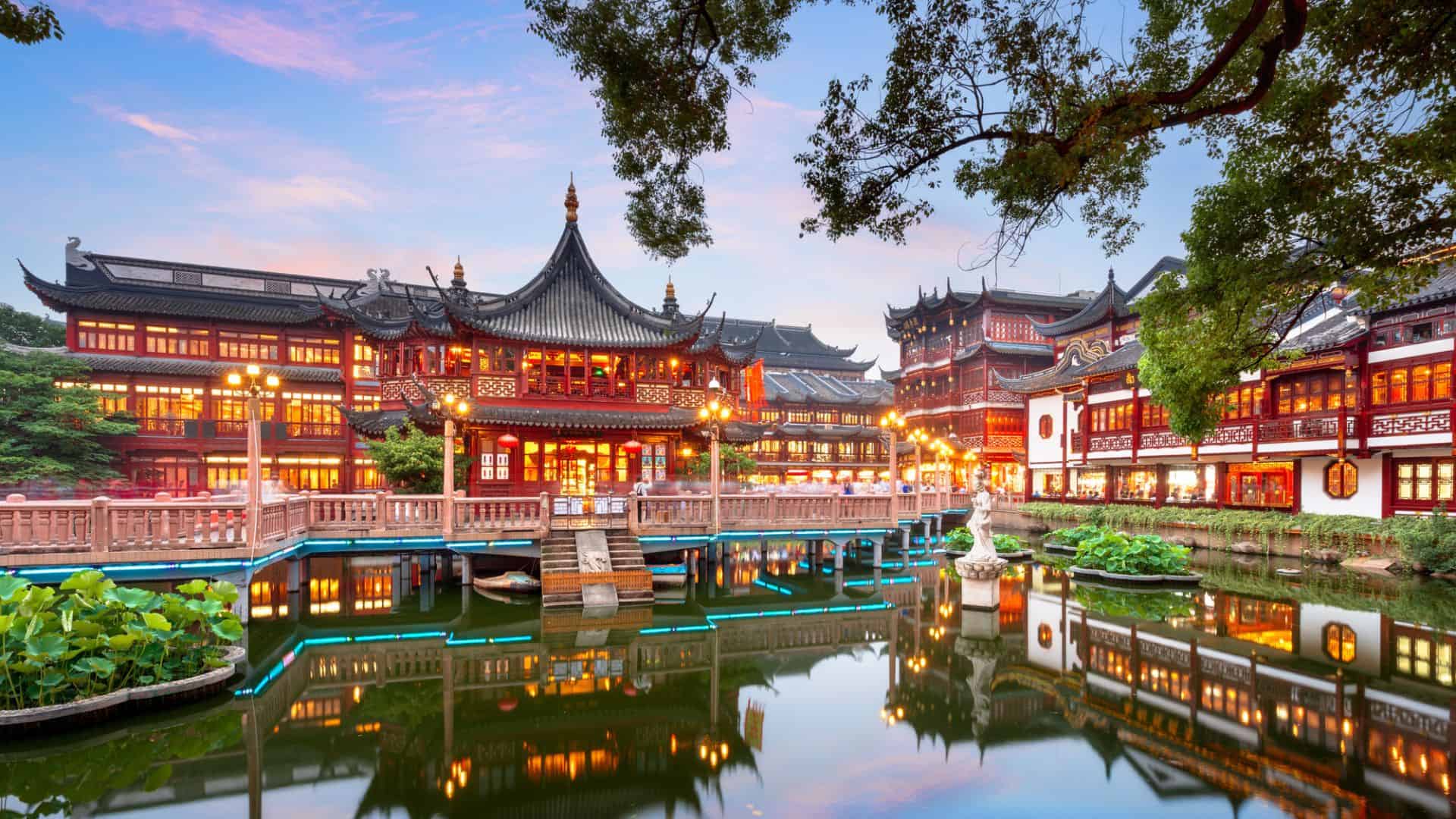 Traditional Chinese buildings with ornate roofs are reflected in a pond at dusk, with trees and lantern lights.