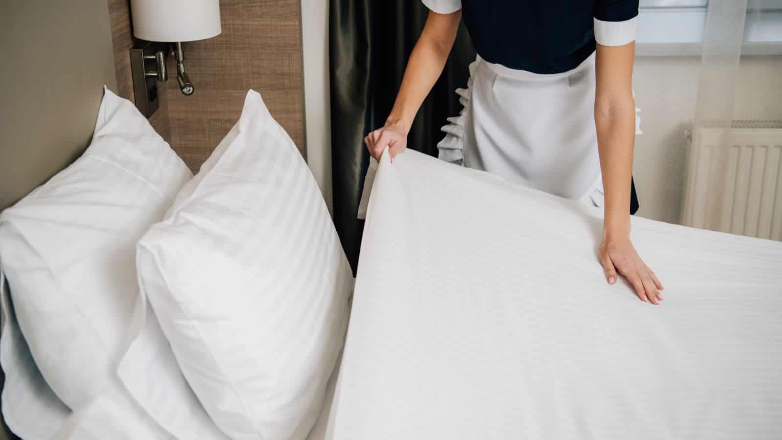 A person in a uniform making a neatly arranged bed with white linens in a hotel room, showcasing one of the travel tips for guests over 35 who value comfort and cleanliness during their hotel booking experience.