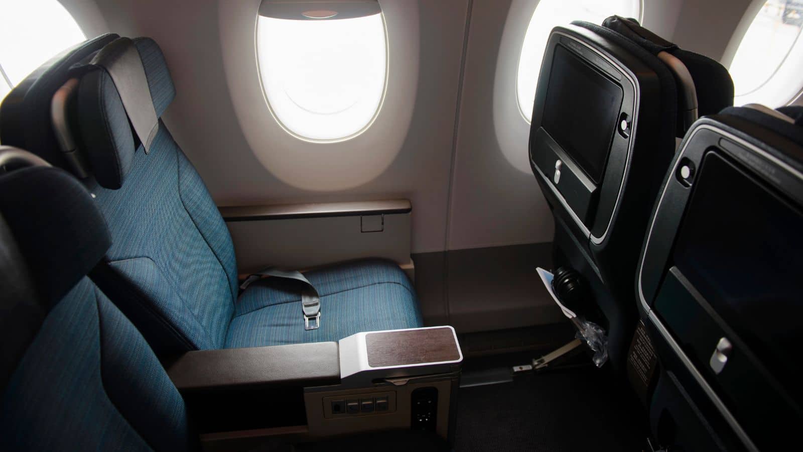 A blue airplane seat by a window with a seatbelt and entertainment screens on the seatbacks ahead.