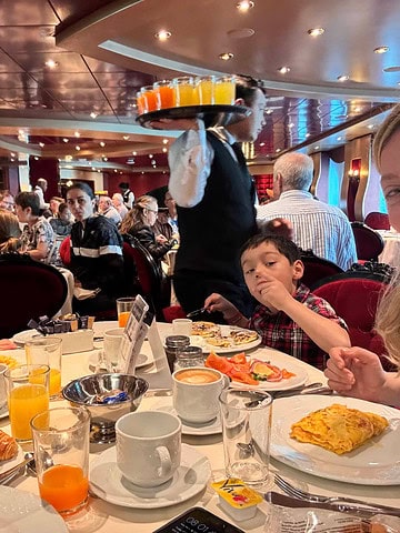 A woman and child enjoy breakfast at a busy restaurant; in the background, a waiter carries a tray of juice, creating a lively cruise dining experience.