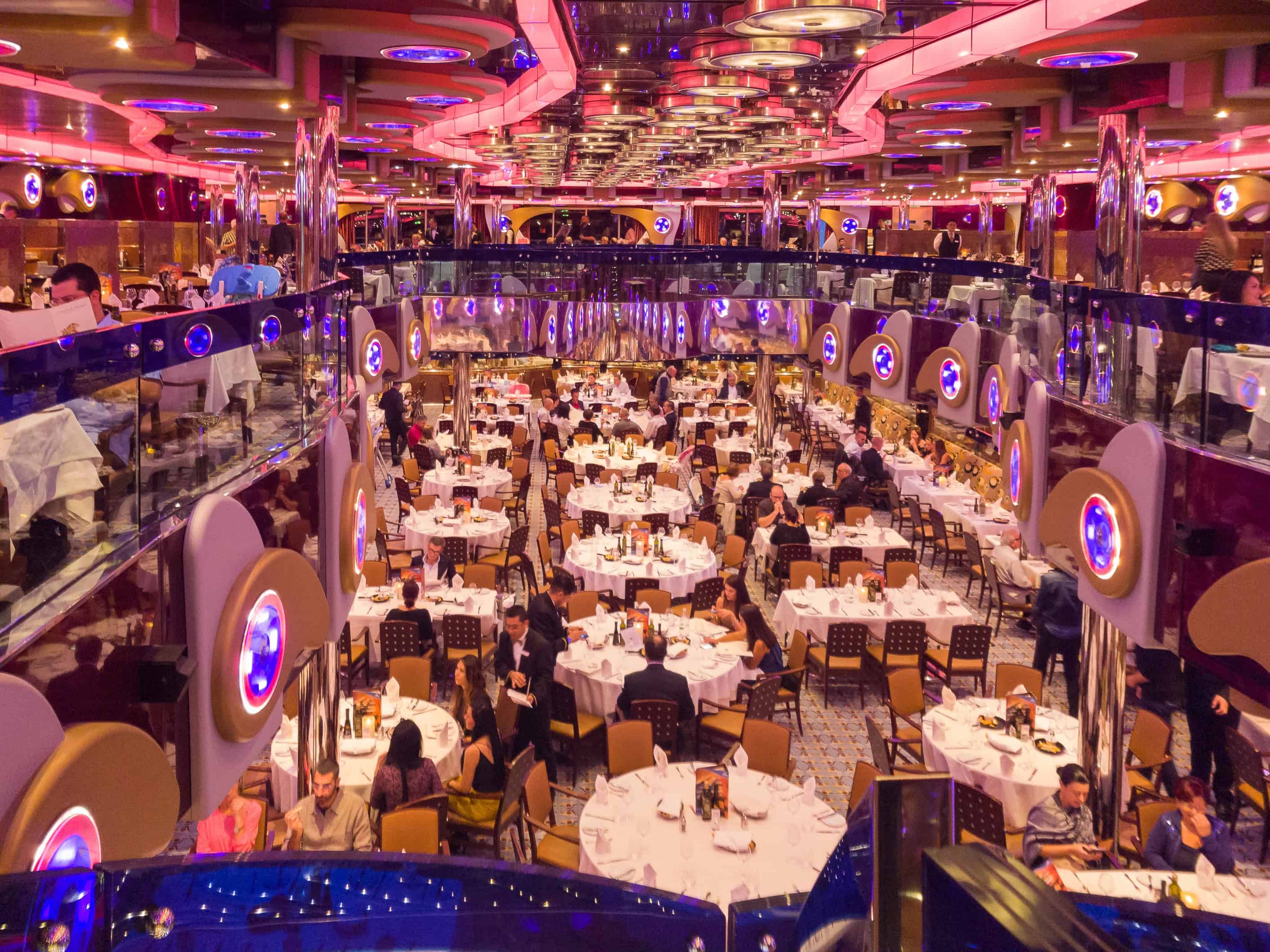 Large, elegant dining hall with round tables, white tablecloths, and many people enjoying cruise ship food while dining under colorful lights.