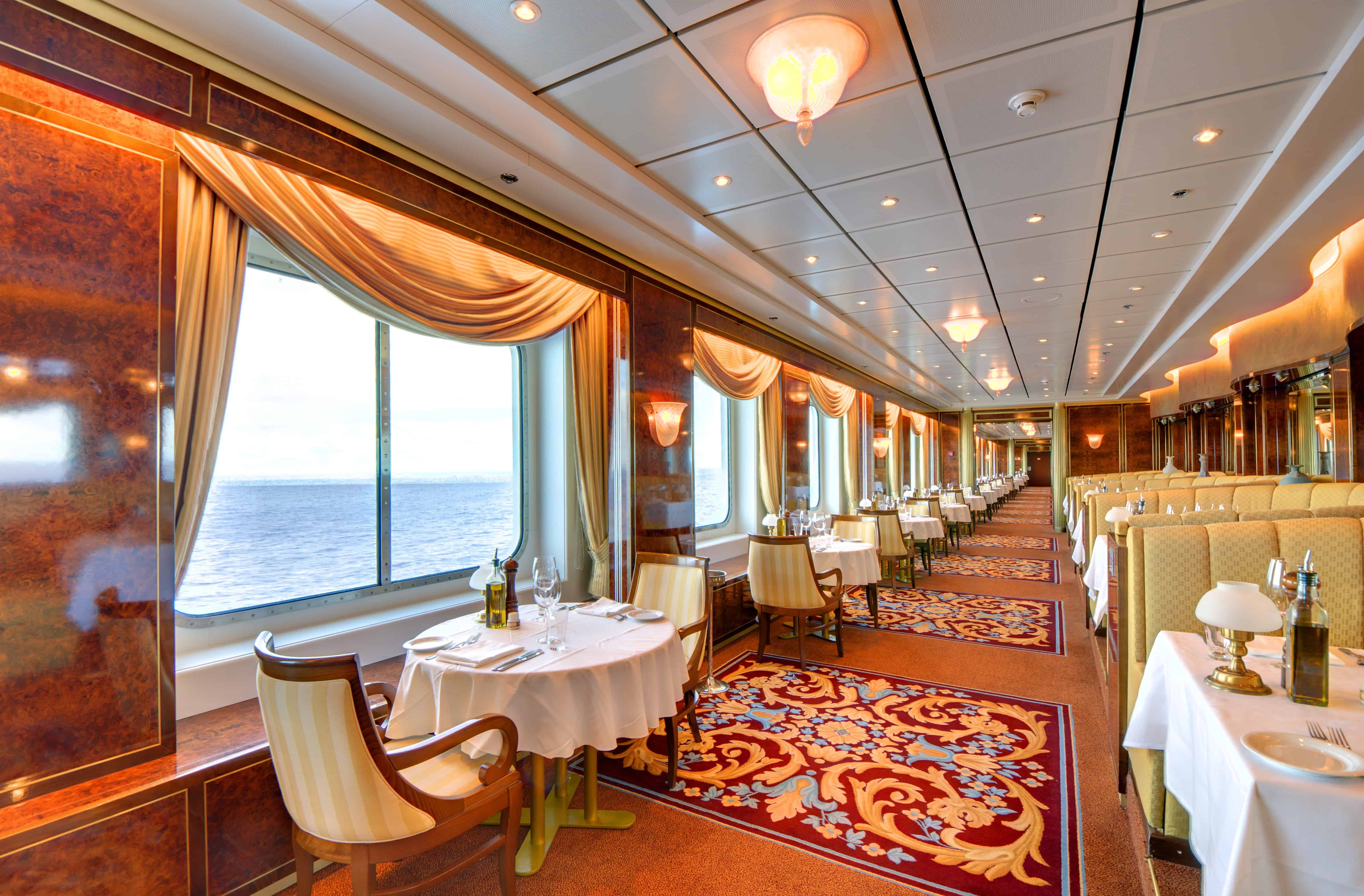 Elegant dining area on a cruise ship with ocean views, set tables, and ornate decor under warm lighting&mdash;perfect for a refined cruise dining experience.