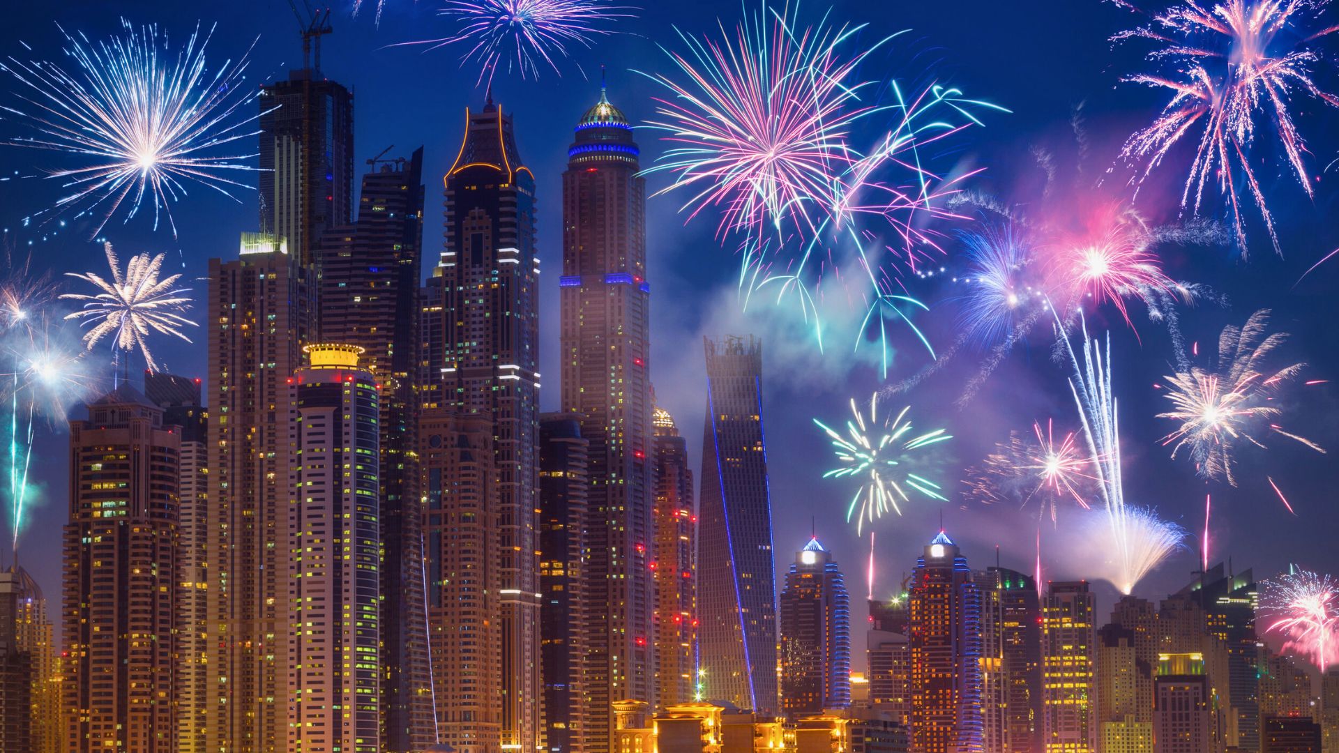 Colorful fireworks light up the night sky above a city skyline filled with tall, illuminated buildings.
