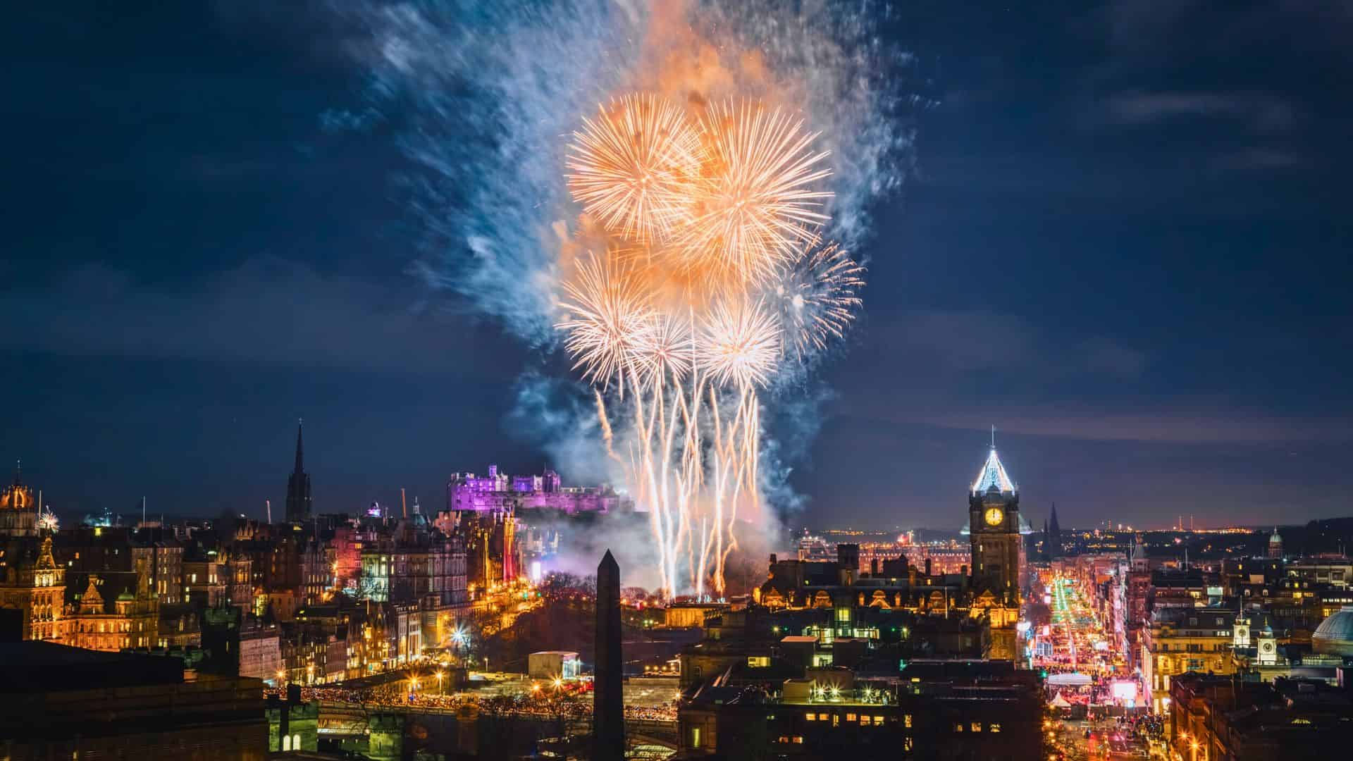 Colorful fireworks light up a city skyline at night, with illuminated buildings and crowded streets below.