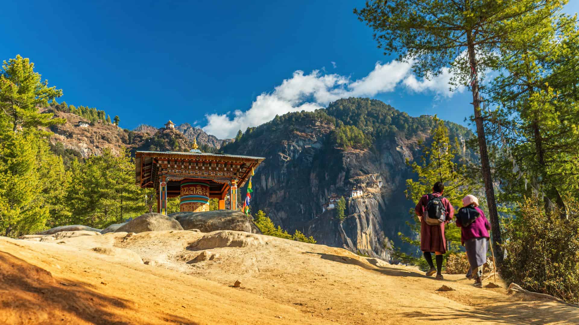 Two hikers walk toward a colorful shrine with mountains and forests in the background under a blue sky.