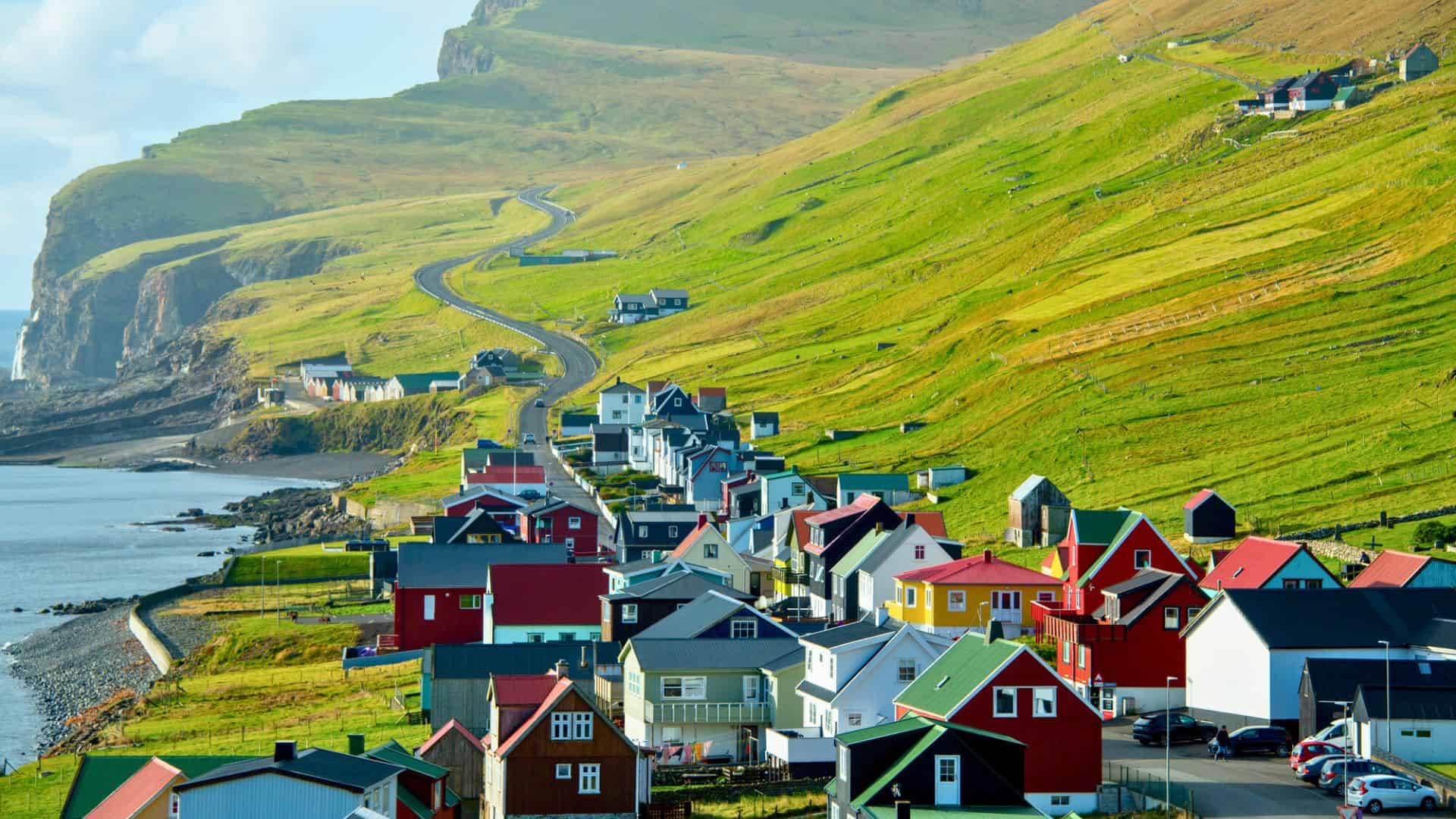 Colorful houses line a coastal village beneath green hills and a curving road by the sea.