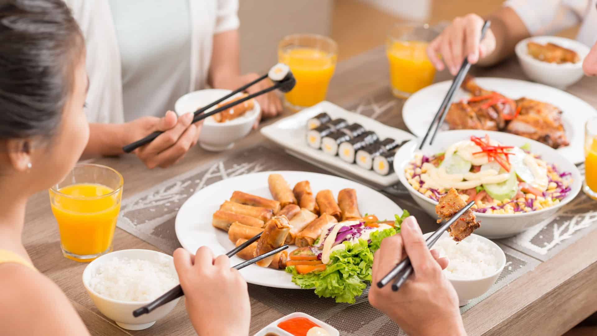 Four people eating Asian food with chopsticks, sushi, spring rolls, rice, salad, and orange juice on the table.