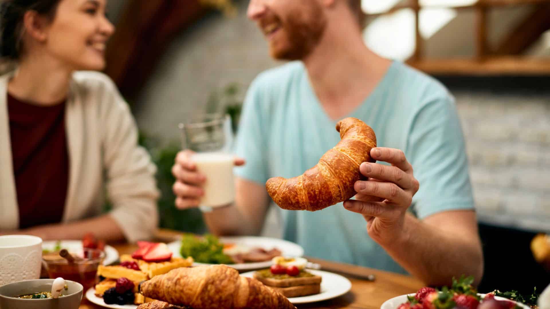 Smiling people enjoy breakfast together; one person holds a croissant and a glass of milk at the table.