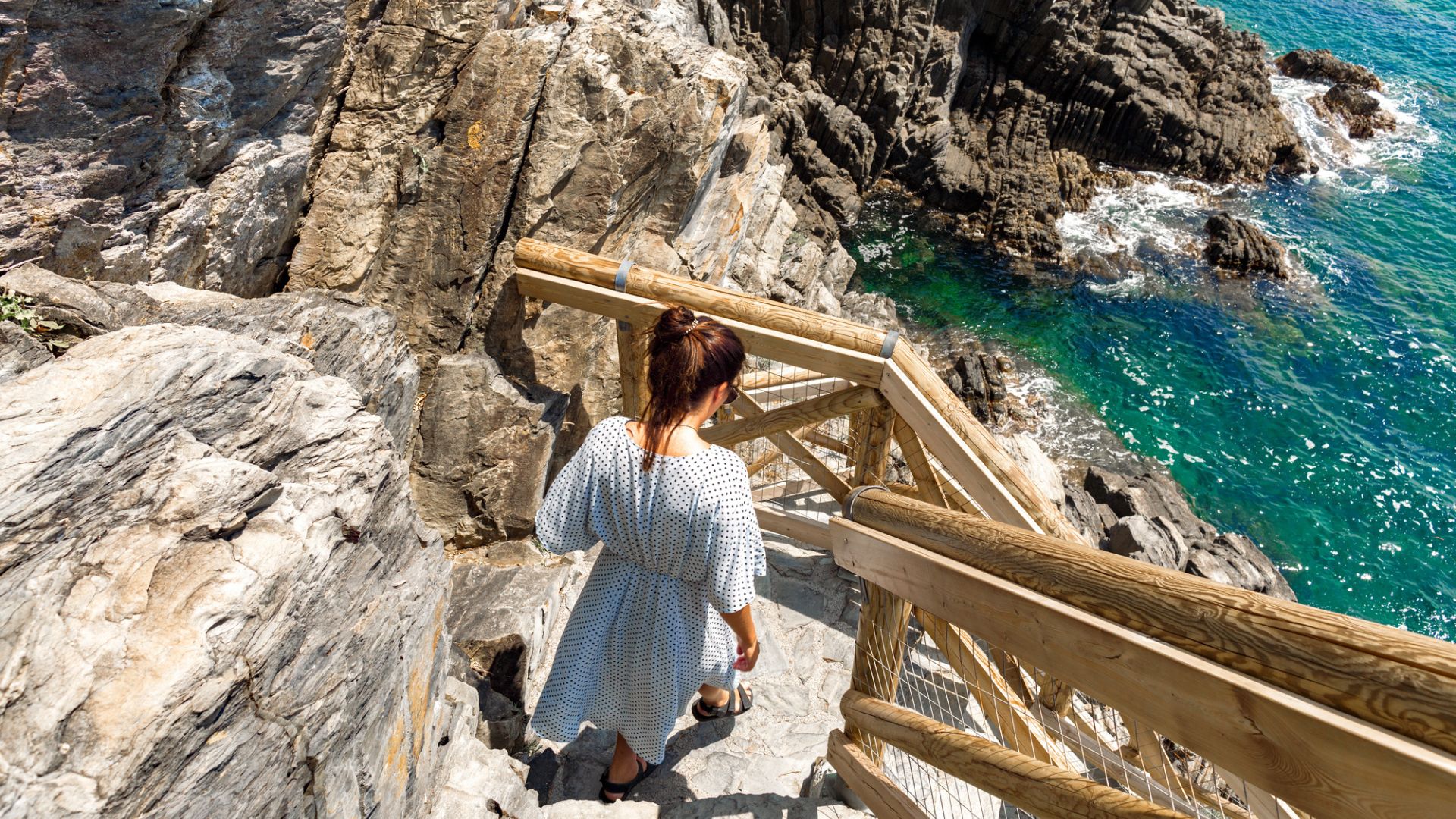 Woman in a white dress walks down wooden stairs along rocky cliffs by the turquoise sea.