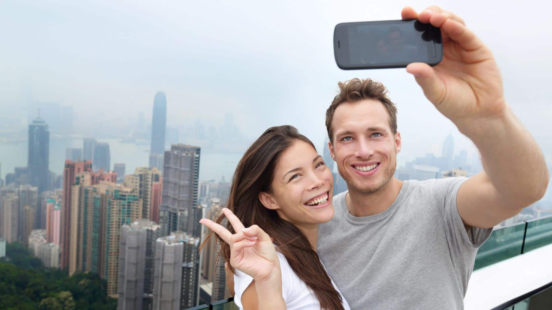 Smiling couple takes a selfie on a rooftop with a city skyline and tall buildings in the background.