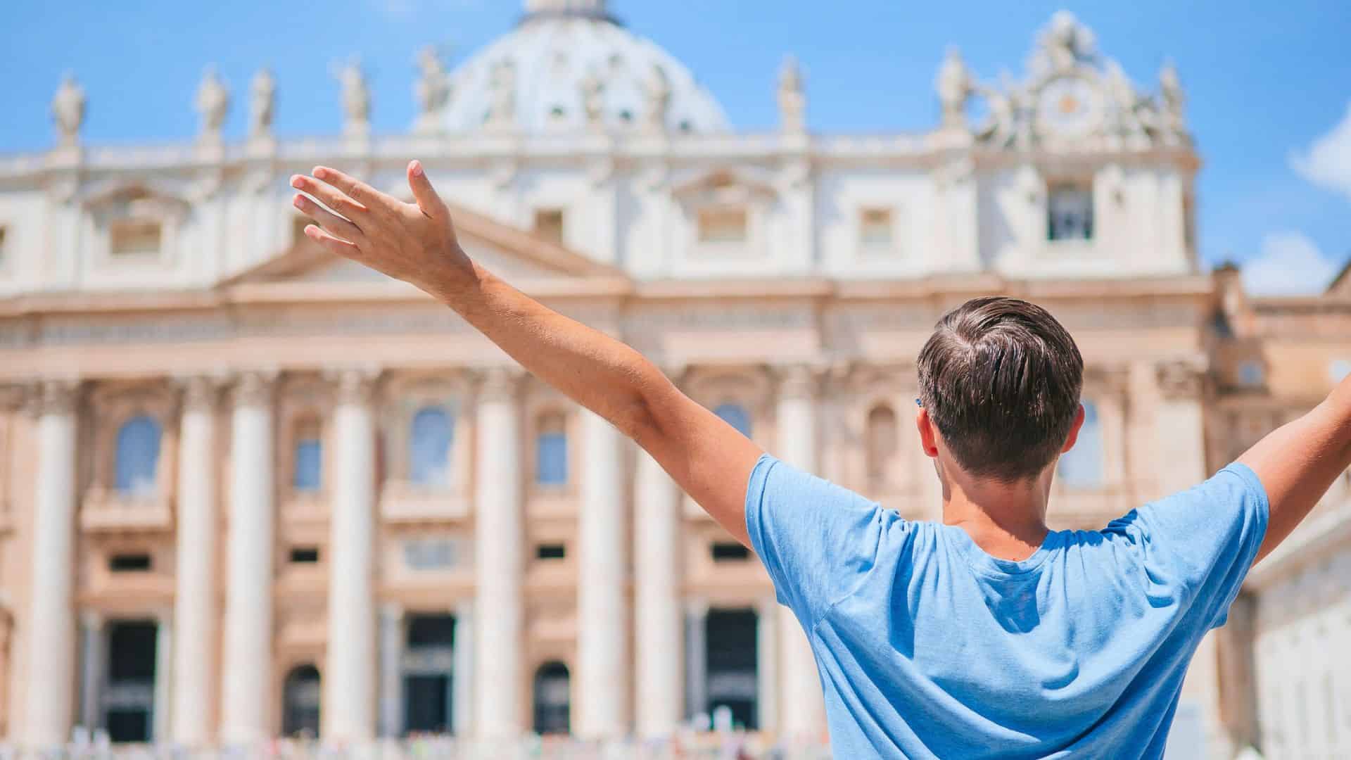 Person in a blue shirt standing with arms raised in front of a large, ornate historic building.