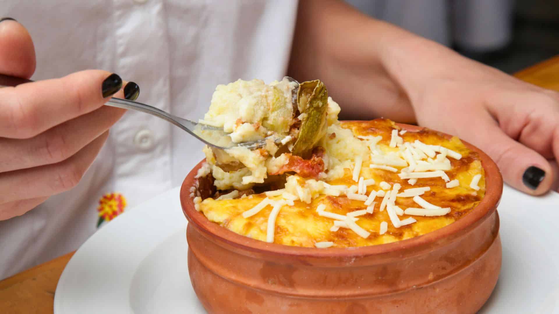 A person holding a forkful of baked casserole topped with melted cheese in a clay dish.