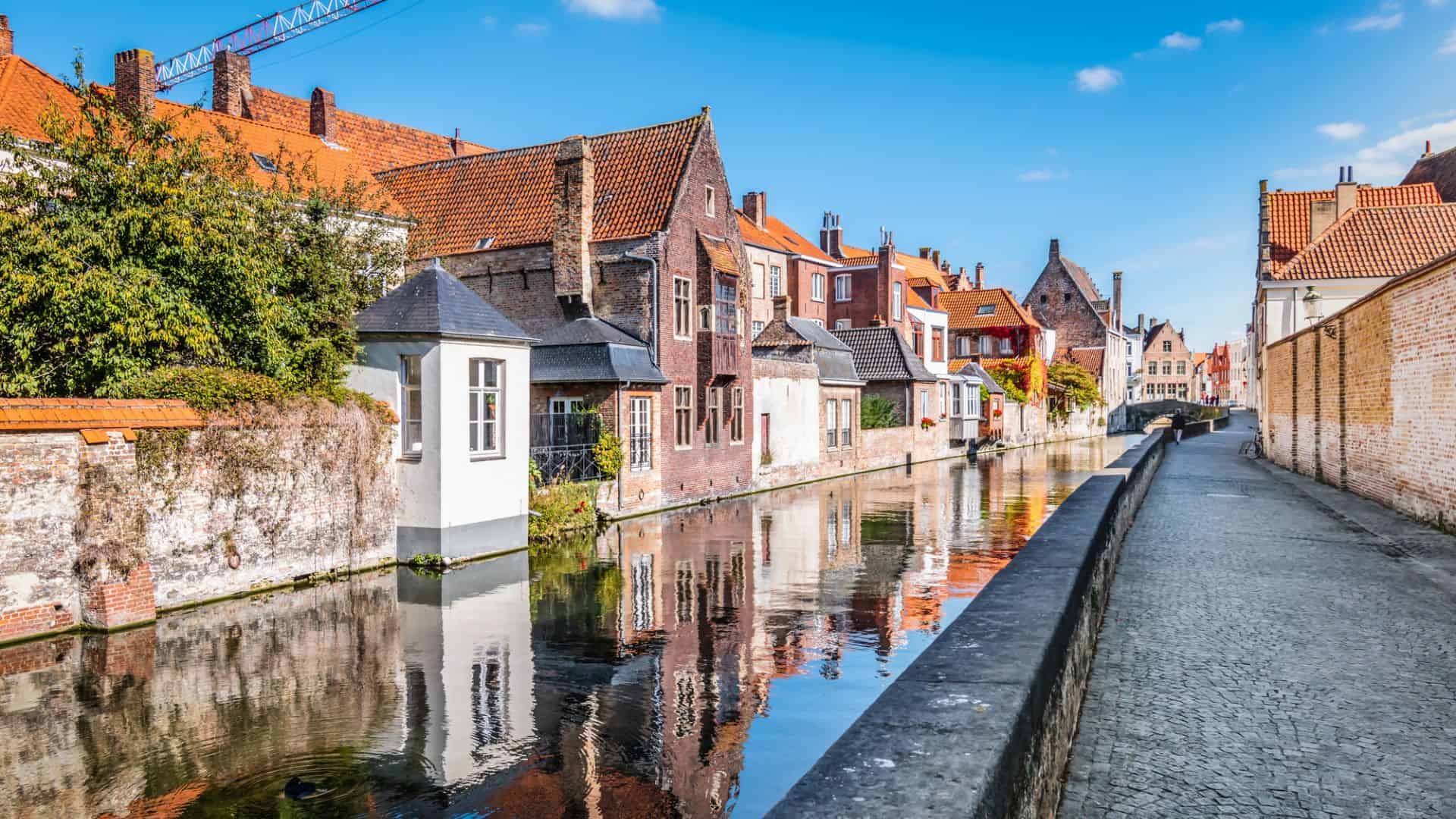 Canal lined with old brick houses and cobblestone path under a clear blue sky in Bruges, Belgium.