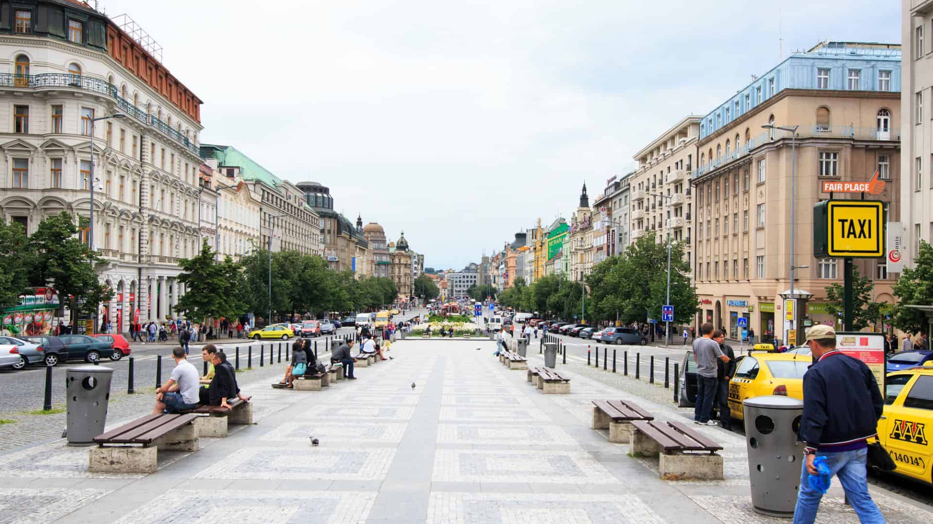 Busy city street with people sitting on benches, cars, and historic buildings lining both sides, taxi sign visible.