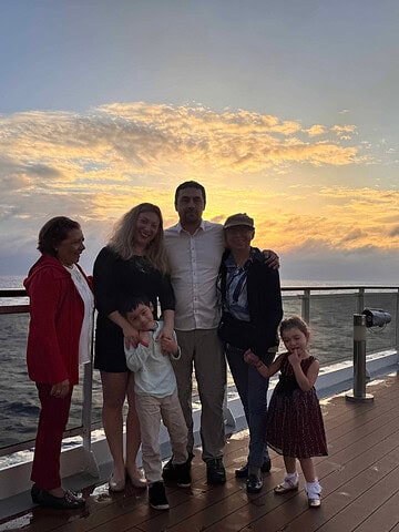 Five adults and two children pose on a ship deck at sunset during their holiday cruise, with the ocean and cloudy sky in the background.