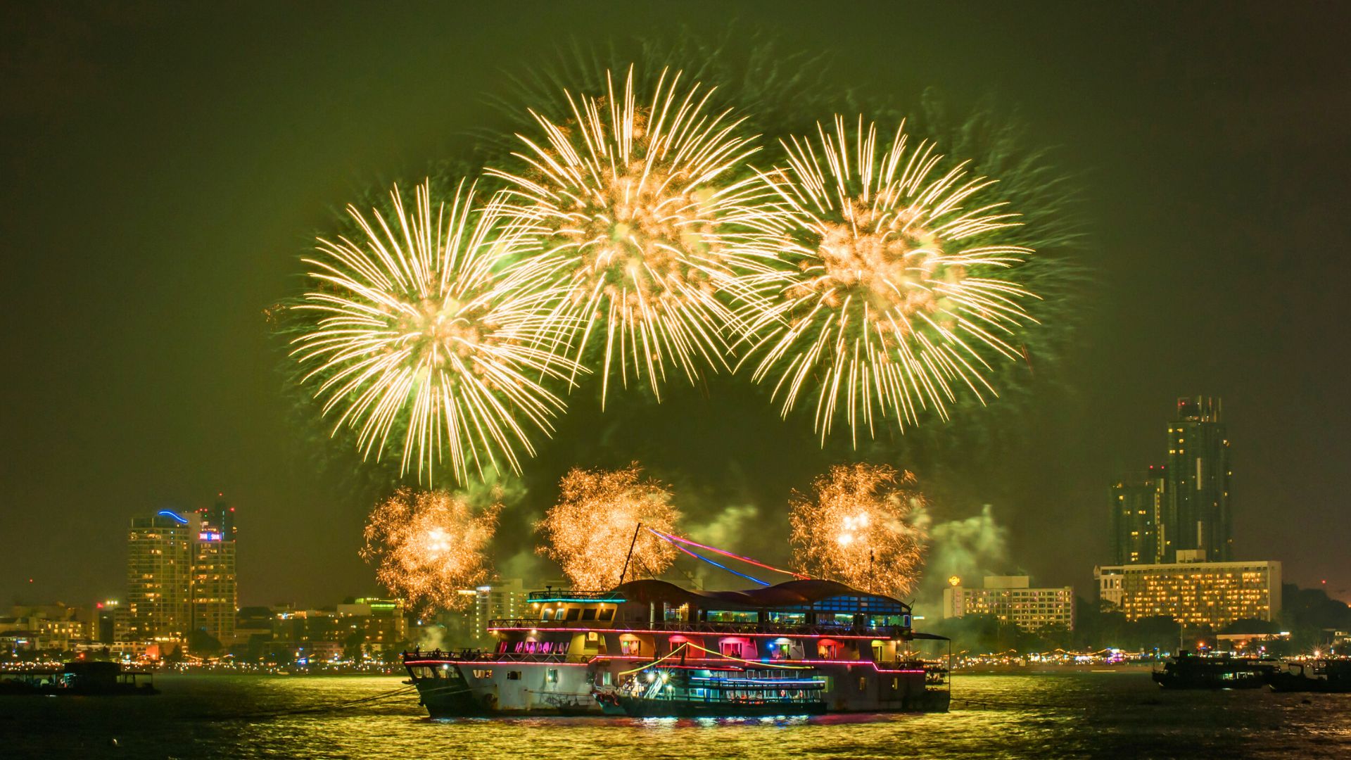 A brightly lit boat on water with colorful fireworks exploding in the night sky above a city skyline.