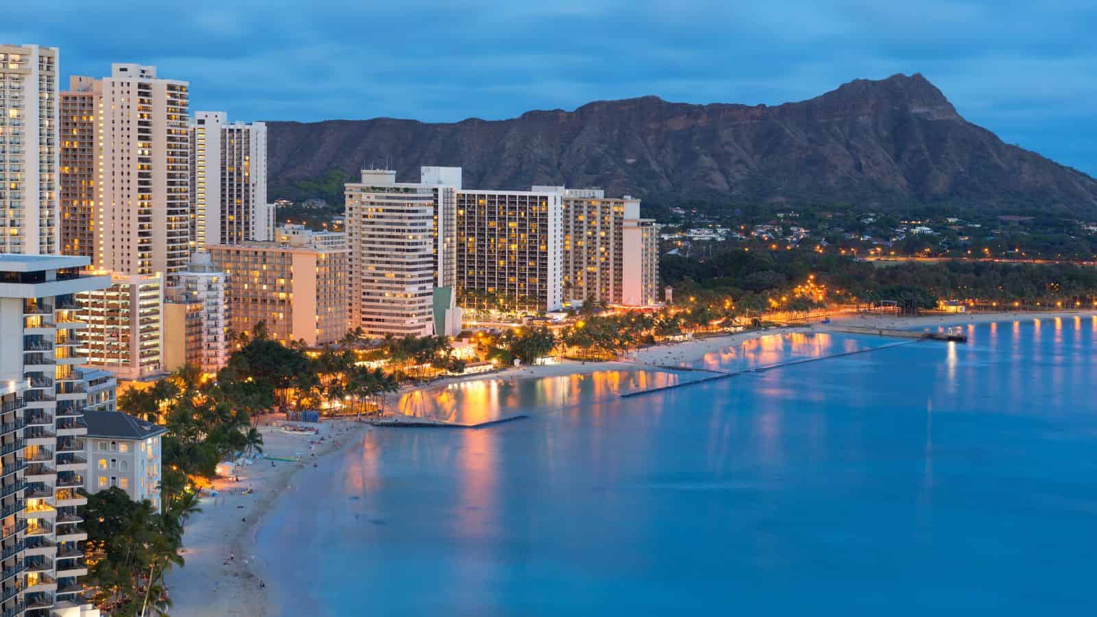 High-rise hotels along Waikiki Beach at dusk, with Diamond Head mountain in the background.