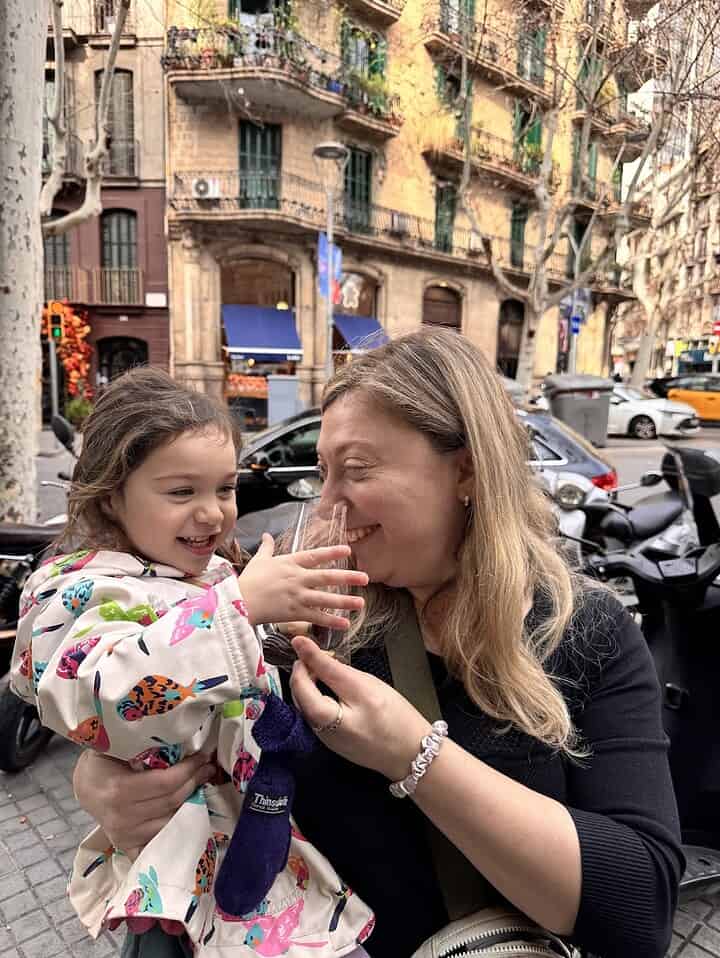 A woman and a smiling girl share a glass cup on a city street with buildings in the background, capturing the joy of spending 4 days in Barcelona together.