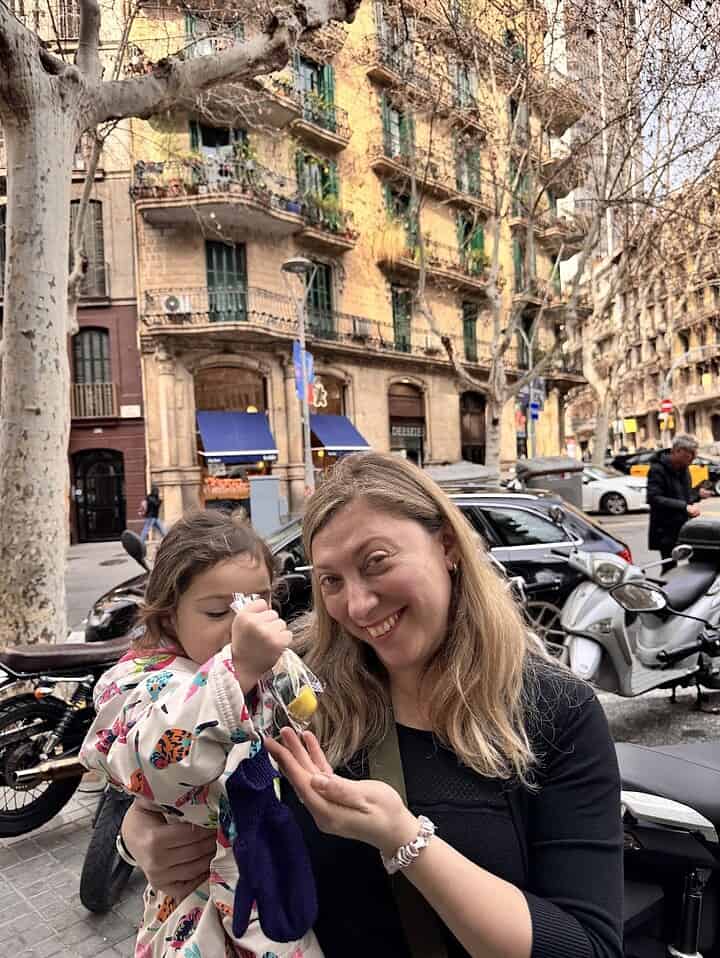 A smiling woman holds out her hand as a young girl pours something into it, capturing a joyful moment during their 4 days in Barcelona, with city buildings in the background.