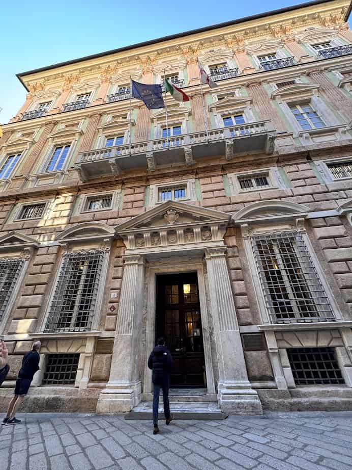 Basking in the sun, a person stands before a grand historic building in Genoa, Italy, its ornate columns and flags capturing the charm of 24 hours in this vibrant city.