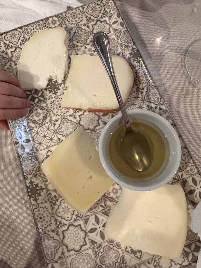 A decorative plate features four cheese slices, a bowl of honey, a spoon, and a hand holding a cheese piece—perfect for savoring the flavors of Genoa, Italy in just 24 hours.
