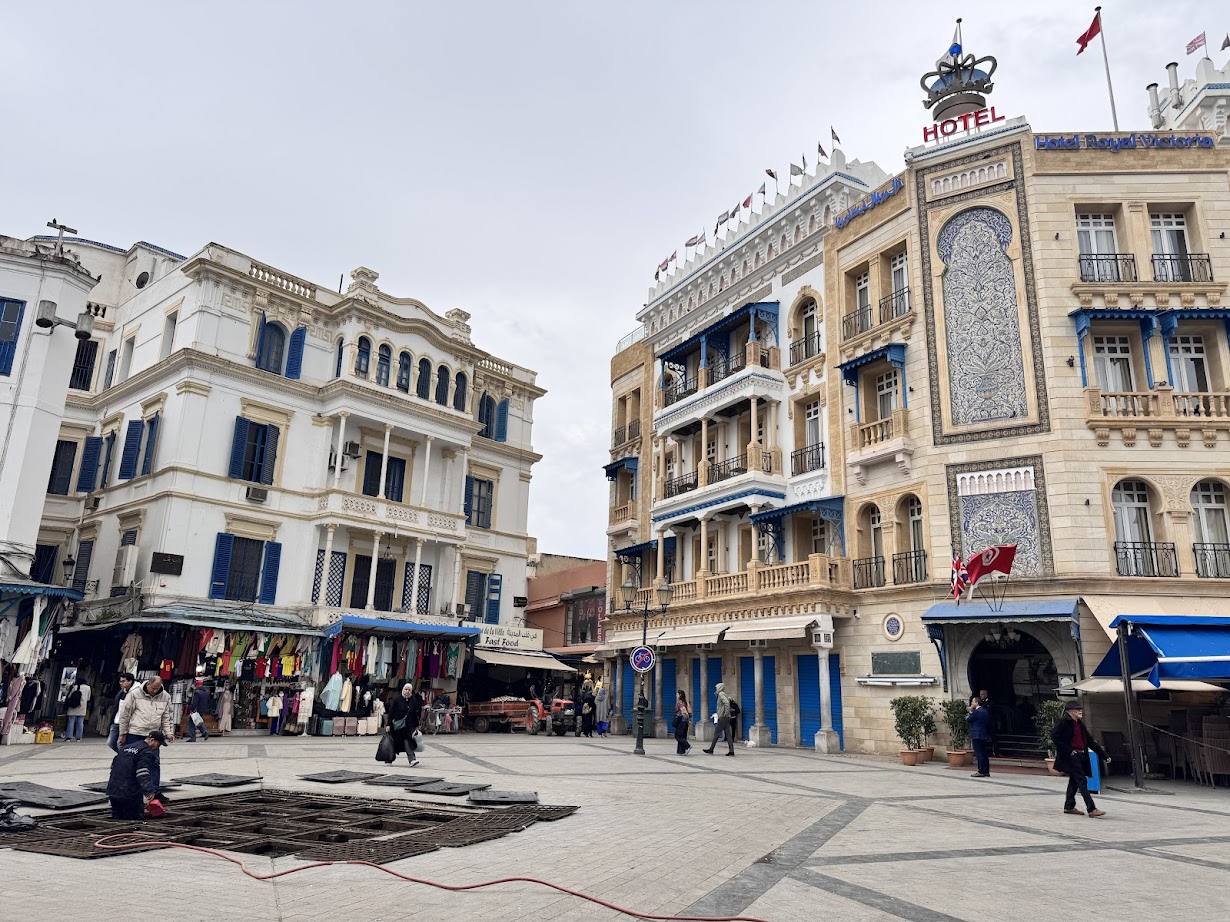 A city square with historic buildings, shops, and people walking on a cloudy day—a perfect stop on your Tunisia itinerary 1 day adventure.
