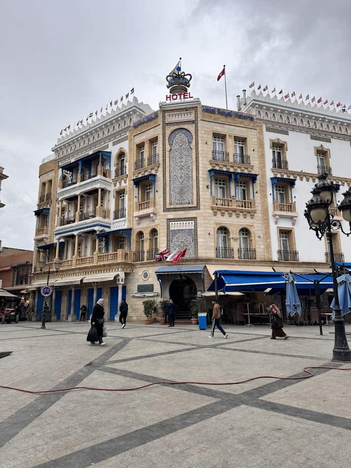 People walk near a historic hotel with blue balconies and many flags under a cloudy sky in a city square—a highlight for any Tunisia itinerary 1 day.