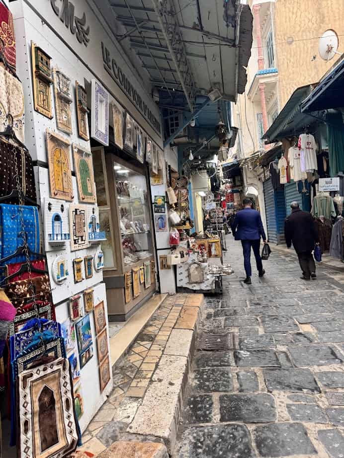 Narrow cobblestone street with shops selling textiles and souvenirs, two people walking away—a perfect scene to include on your Tunisia itinerary 1 day adventure.