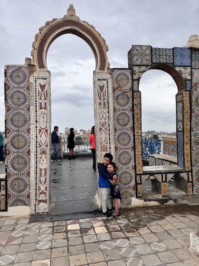 Two children hugging and smiling under a colorful tiled archway, with people and a city view in the background—a heartwarming scene to spot on your Tunisia itinerary 1 day adventure.