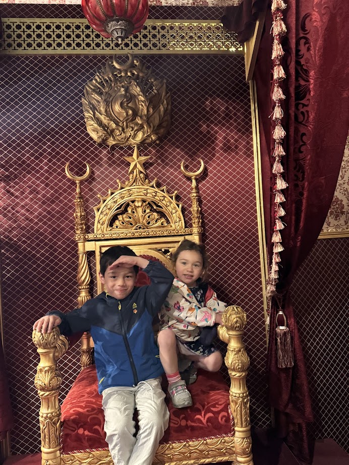 Two children sit on an ornate golden throne with red velvet upholstery and rich red drapery behind them, evoking the regal splendor you might discover on a Tunisia itinerary 1 day adventure.