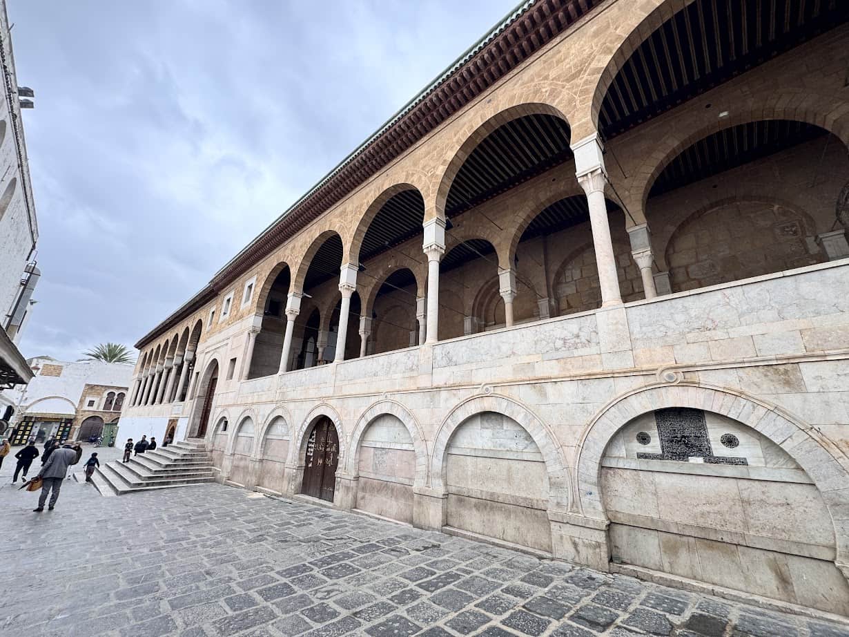 Historic stone building with arched columns and walkways, people strolling on a cobblestone courtyard under a cloudy sky—an ideal scene to include in your Tunisia itinerary 1 day adventure.