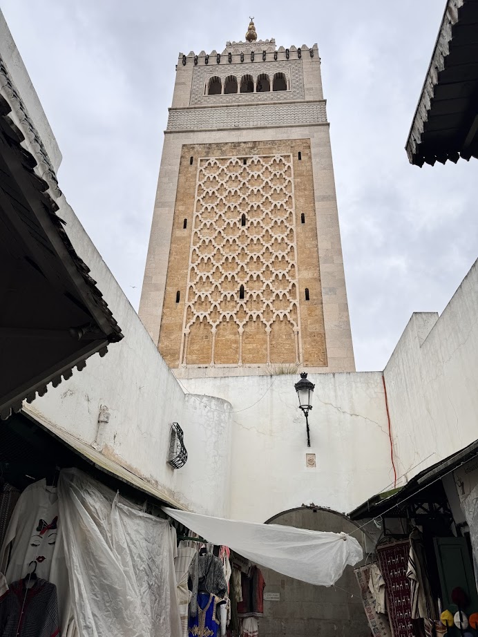 Tall ornate minaret rises above market stalls and whitewashed walls under a cloudy sky—a must-see highlight on any Tunisia itinerary 1 day.