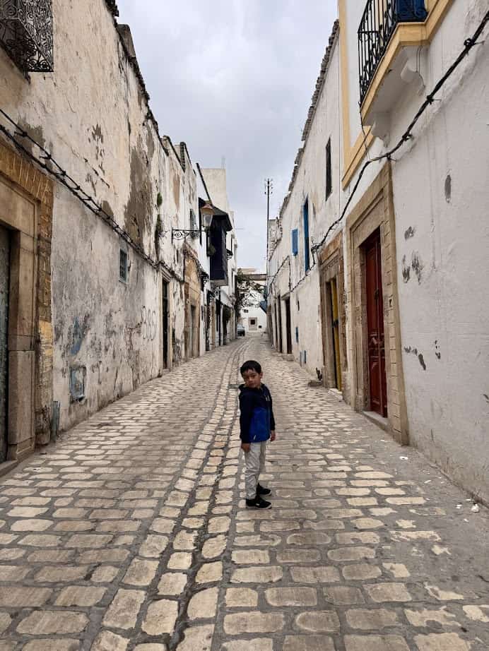 A young boy stands on a narrow cobblestone street lined with old, weathered buildings, capturing the charm you might find on a Tunisia itinerary 1 day adventure.