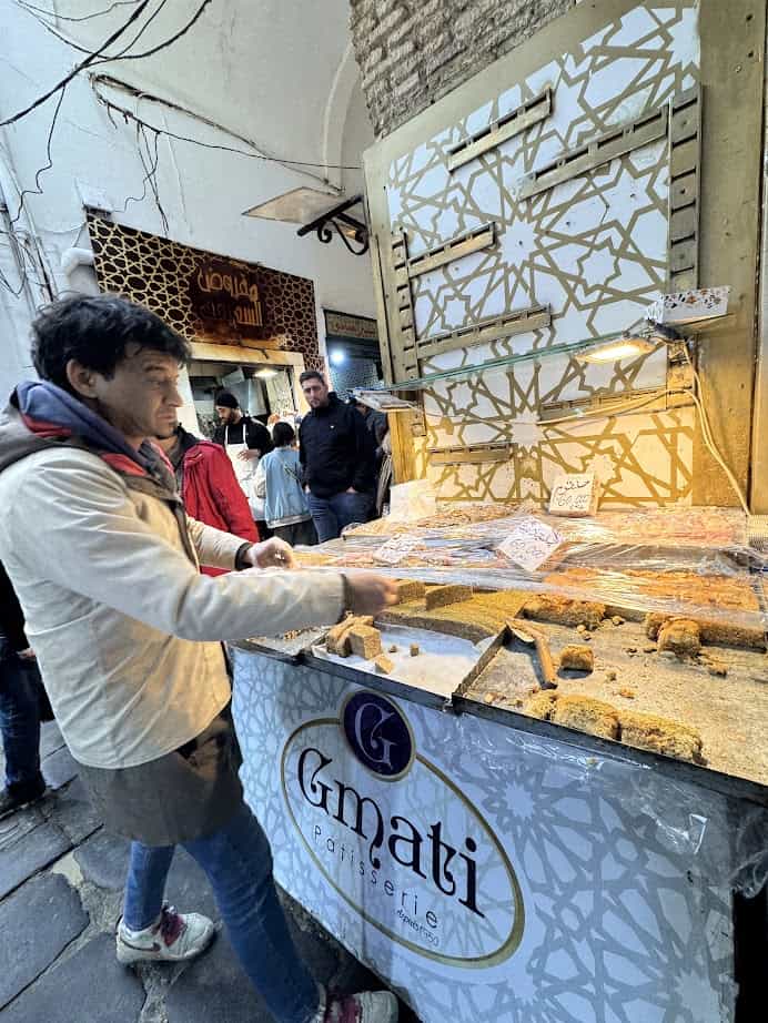 A vendor slices sweets at a street stall with people standing nearby in a bustling market—an authentic moment to include in your Tunisia itinerary 1 day adventure.