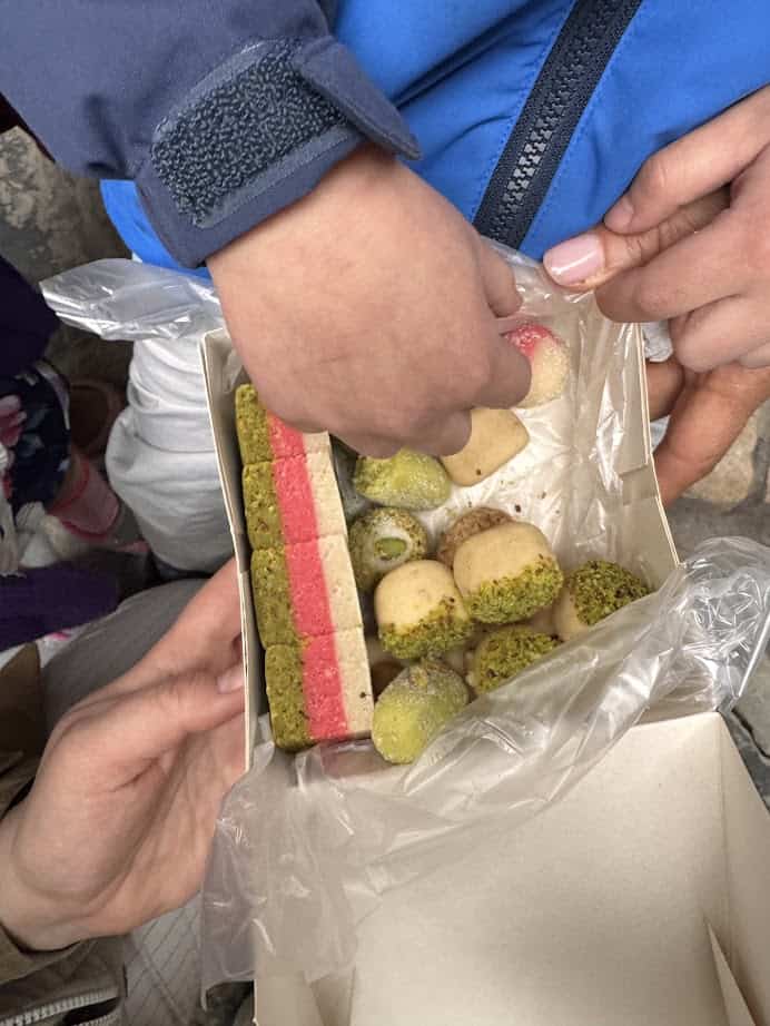 A child reaches into a box of colorful assorted sweets, some coated in green pistachio crumbs—a delightful treat to savor during your Tunisia itinerary 1 day adventure.