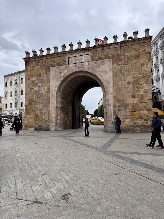 Stone city gate with arched entrance, people walking nearby, and Tunisian flags on top under a cloudy sky—a perfect stop on your Tunisia itinerary 1 day adventure.