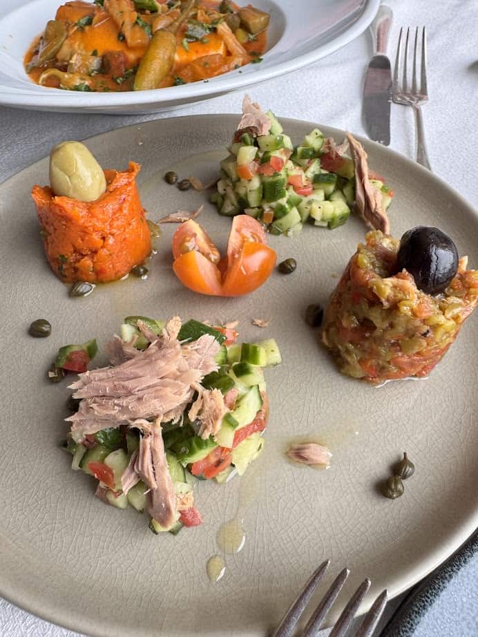 A plate with assorted vegetable salads, tuna, olives, capers, and a tomato garnish evokes the fresh flavors you might enjoy on a Tunisia itinerary 1 day adventure, with pasta in the background adding to the Mediterranean feast.