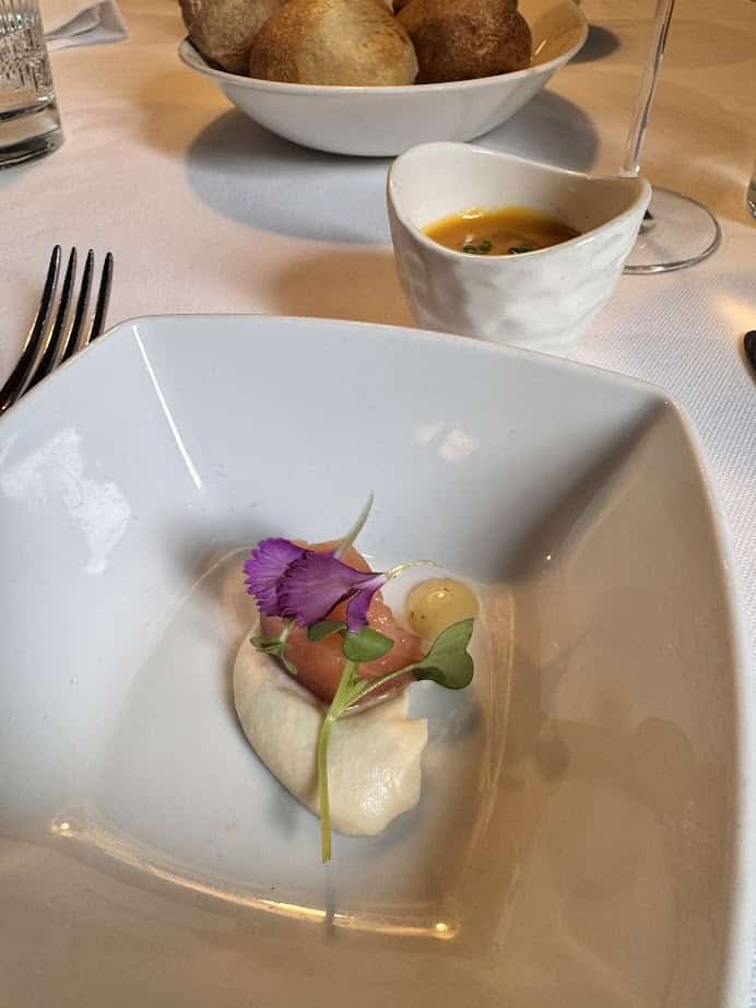 A gourmet appetizer with garnish in a white bowl, set on a table with bread and soup in the background&mdash;perfect for savoring during your best Barcelona trip.