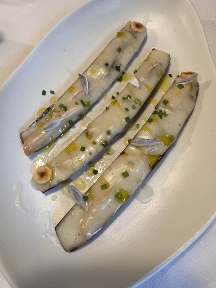 Savor three razor clams on a white plate, topped with melted cheese, chives, and a drizzle of oil&mdash;a must-try dish for any Barcelona travel foodie during your 4 days in Barcelona.