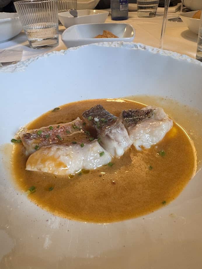 Three pieces of fish in an orange broth, garnished with chives and served in a white bowl&mdash;perfect for those planning their Barcelona itinerary or seeking flavors to try during 4 days in Barcelona.