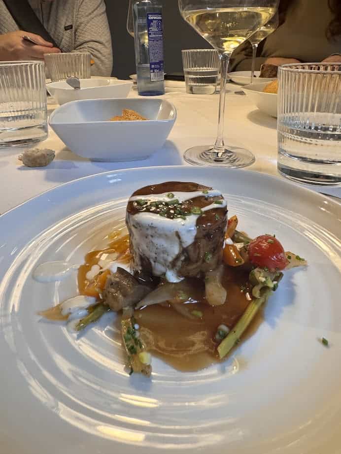 A gourmet dish with vegetables and sauce on a white plate, set on a table with glasses and a wine glass&mdash;perfect for savoring during your 4 days Barcelona adventure or adding to your Barcelona itinerary of culinary delights.
