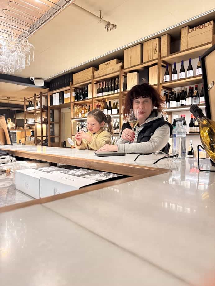 A woman and a girl sit at a bar in Barcelona, each holding a glass, surrounded by shelves of wine bottles and boxes&mdash;an inviting spot to add to your 4 days itinerary for the best things to do in the city.